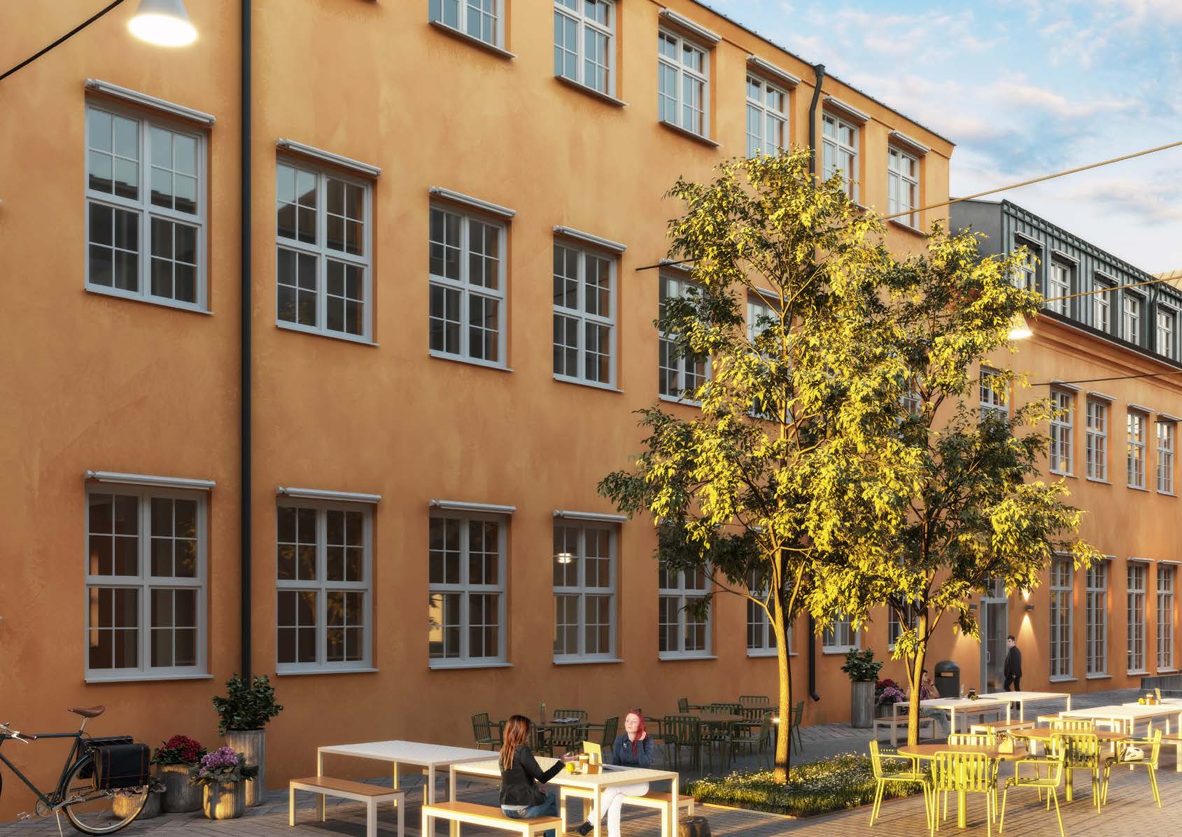 Gård mellan putsade fabriksbyggnader i ockra, med spröjsade fönster i tre rader. Två unga träd står i en plantering mitt på den stenlagda ytan. Vid fasaden står vita bord med bänkar och gula stolar, och två personer sitter vid ett av borden. Längre bort syns en byggnad med glasat sågtandstak.