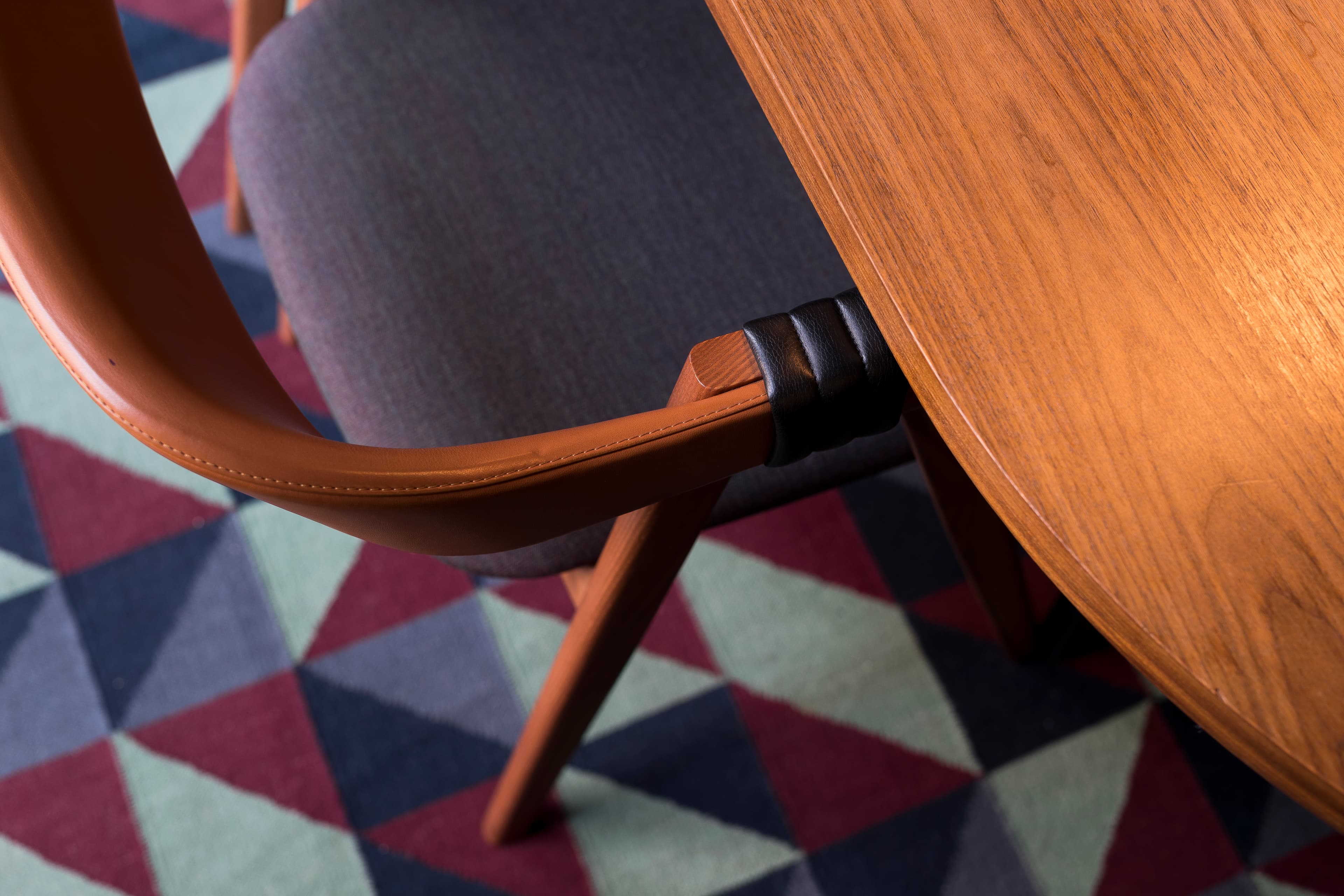 Närbild på en stol i trä med armstöd i brunt läder, intill en bordsskiva i teak. Golvet har en mönstrad matta i geometriska trianglar.