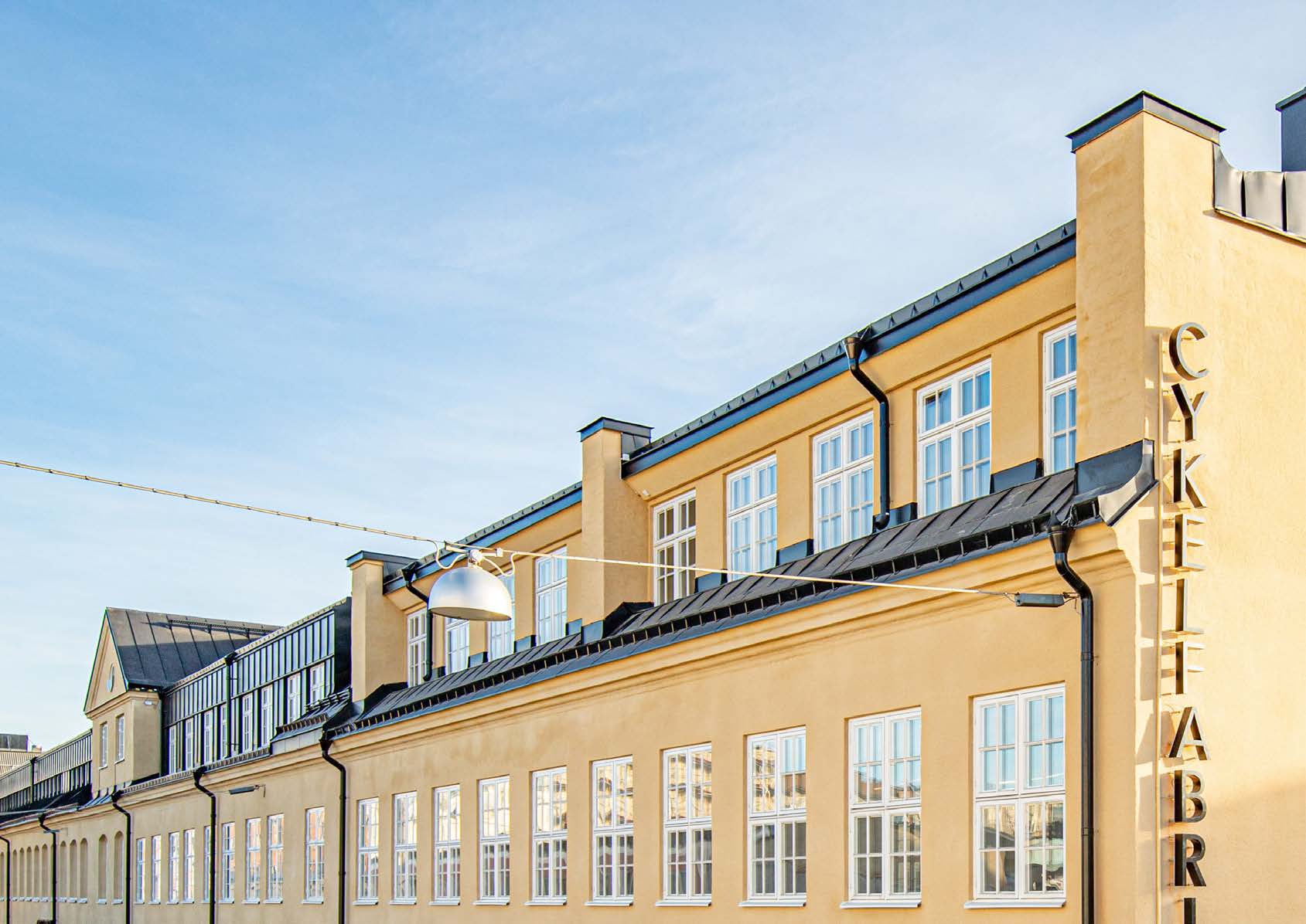 En lång gul putsad fasad i två våningar med en tät rad spröjsade fönster och svart plåttak. På gaveln står CYKELFABRIKEN i stora bokstäver lodrätt nerför hörnet. En rund plåtlampa hänger i en vajer tvärs över gatan.